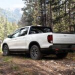 A white 2024 Honda Ridgeline TrailSport is shown on a tree-lined path after leaving a Honda dealer.