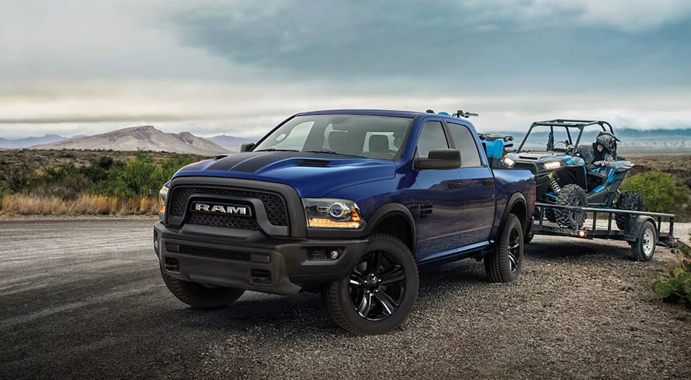 A blue 2025 Ram 1500 Classic Warlock towing an ATV over a dirt road.