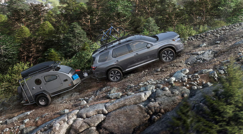 A grey 2025 Honda Pilot TrailSport towing a small camper up a rocky trail.