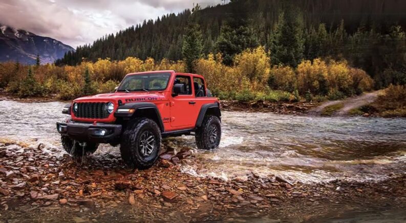 A red 2024 Jeep Wrangler Rubicon parked on a shallow river shore.