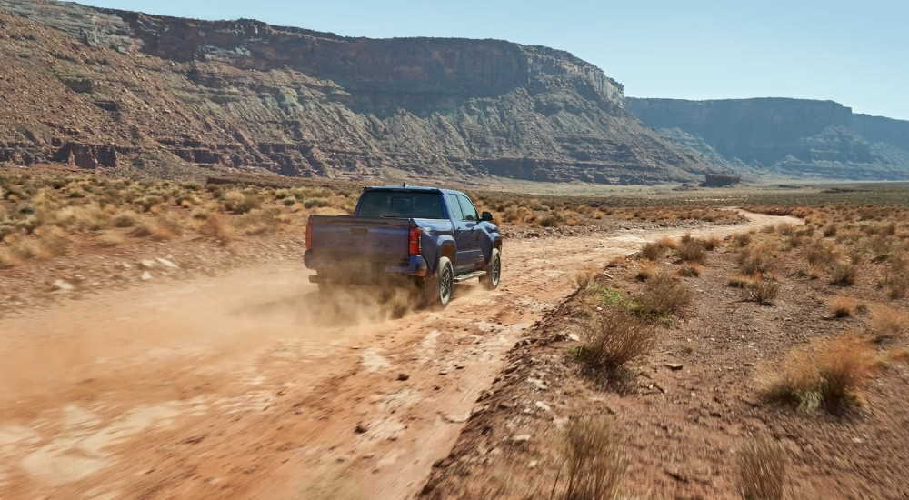 A blue 2024 Toyota Tacoma TRD Sport is show from the rear on a dusty trail.