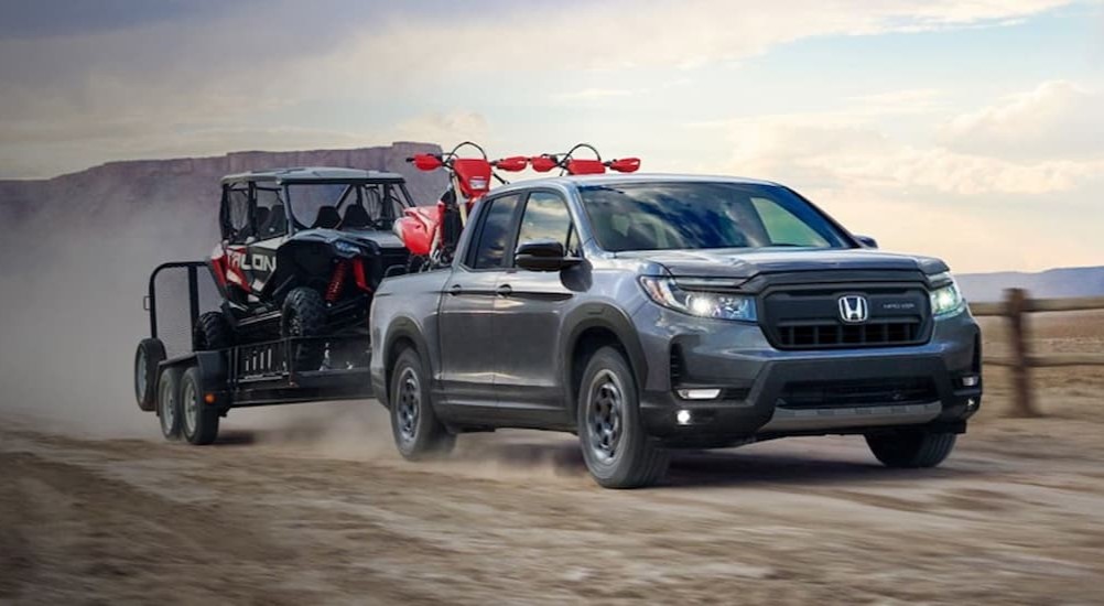 A gray 2024 Honda Ridgeline TrailSport is shown towing a trailer on a dirt road.