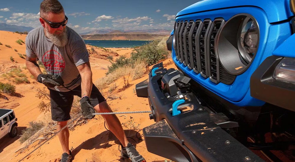 A person is shown using a winch on a blue 2025 Jeep Wrangler.