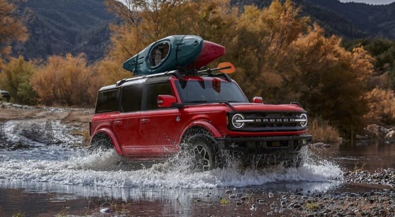 A red 2024 Ford Bronco Outer Banks with a canoe on a roof rack driving through a shallow river.