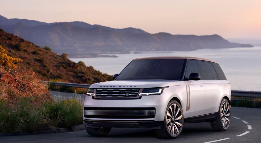 A silver 2026 Range Rover SV shown driving a coastline at sunrise.