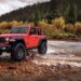 A red 2024 Jeep Wrangler Rubicon parked on a shallow river shore.